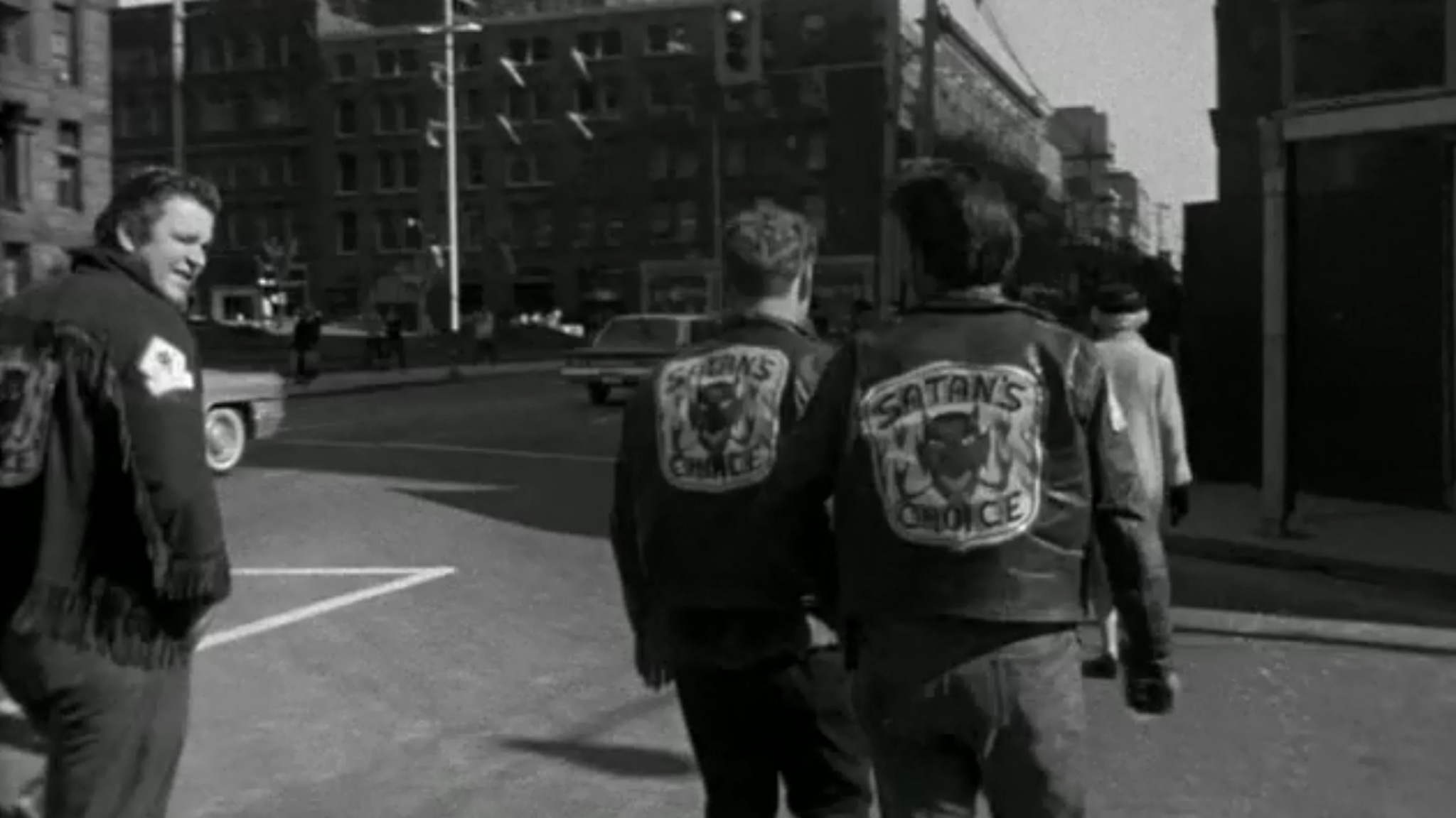 Black-and-white film still of people crossing a city street in leather jackets, two bearing Satan’s Choice back patches.